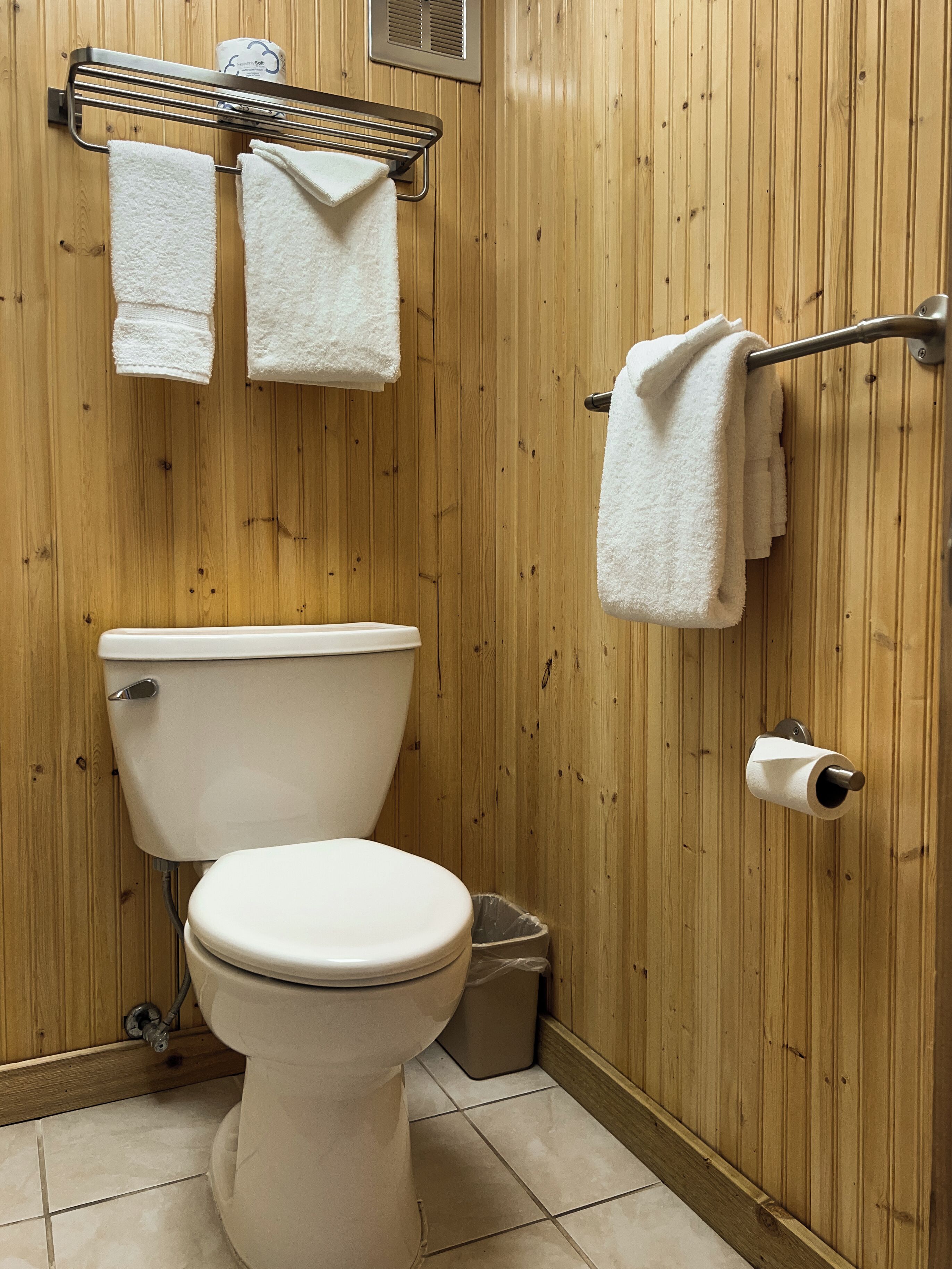 A bathroom with a wooden wall, a toilet, a towel rack with two towels, and a toilet paper holder.