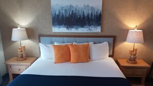 A neatly made bed with white and orange pillows, flanked by wooden nightstands with lamps, in a room with a painting of a forest on the wall. - The Cliff Dweller on Lake Superior (Tofte)