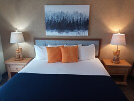 Una cama bien tendida con almohadas blancas y anaranjadas, flanqueada por mesitas de noche de madera con lámparas, en una habitación con un cuadro de un bosque en la pared.