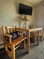 A wooden chair with a cushion, a table, a lamp, and a TV mounted on the wall.