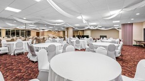 Uma sala de banquetes com mesas redondas cobertas por toalhas brancas e cadeiras, piso carpeteado e teto com iluminação embutida.