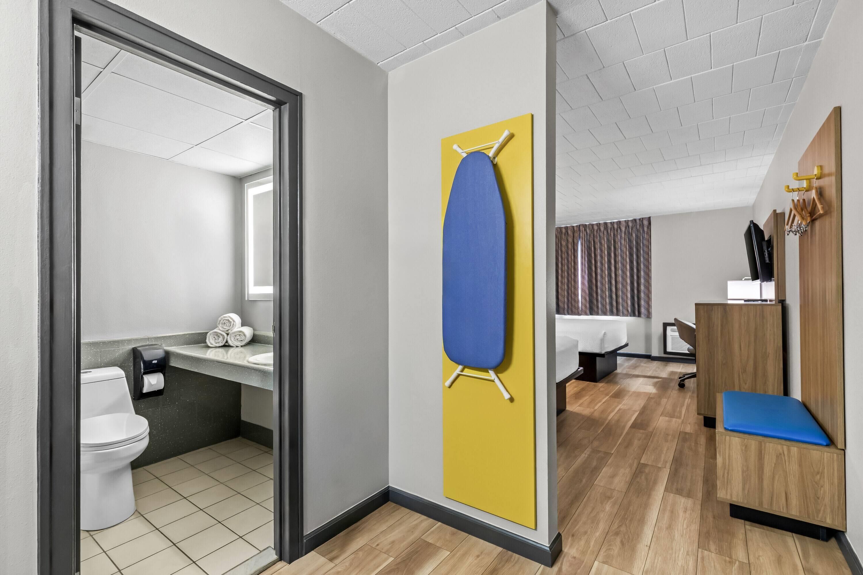 Una habitación de hotel moderna con suelo de madera, una pared de acento amarilla, una tabla de planchar azul, un banco de madera, un escritorio y un baño con inodoro y lavamanos.
