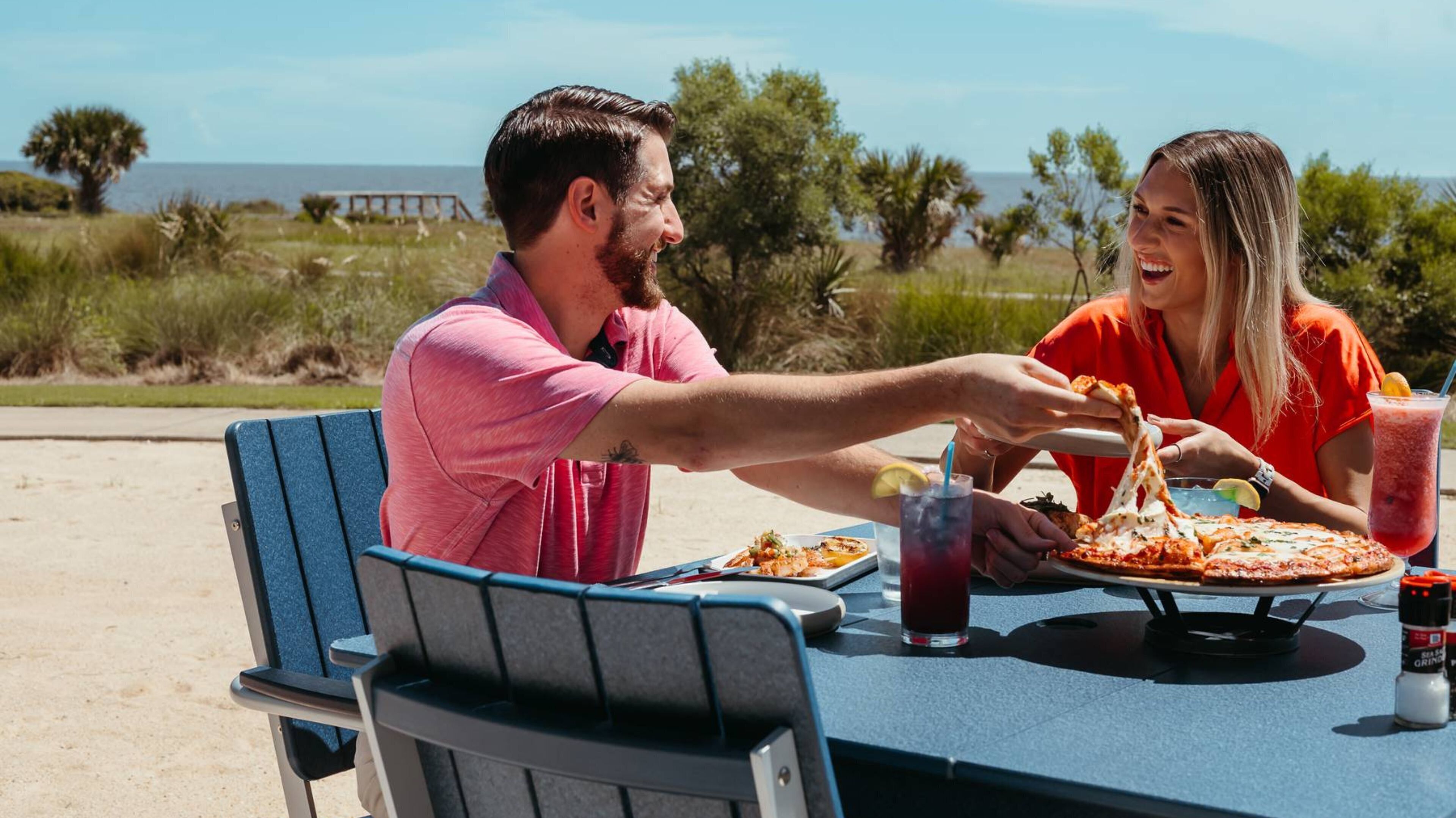Deux personnes sont assises à une table, devant une pizza et des boissons, à l’extérieur, avec vue sur la plage et l’océan.