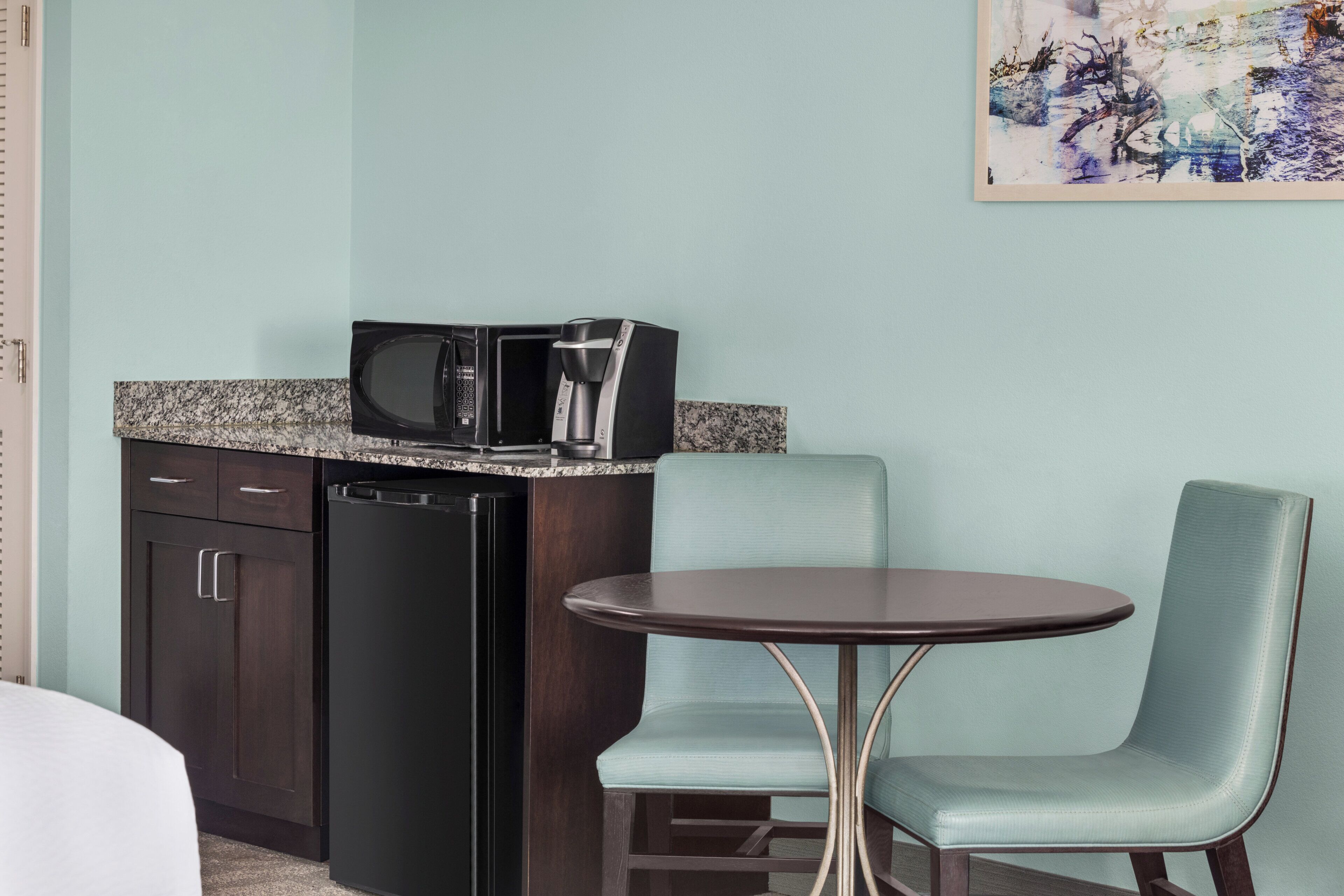 A compact kitchenette with a microwave, coffee maker, and dishwasher. There is a small round table with a chair beside it.