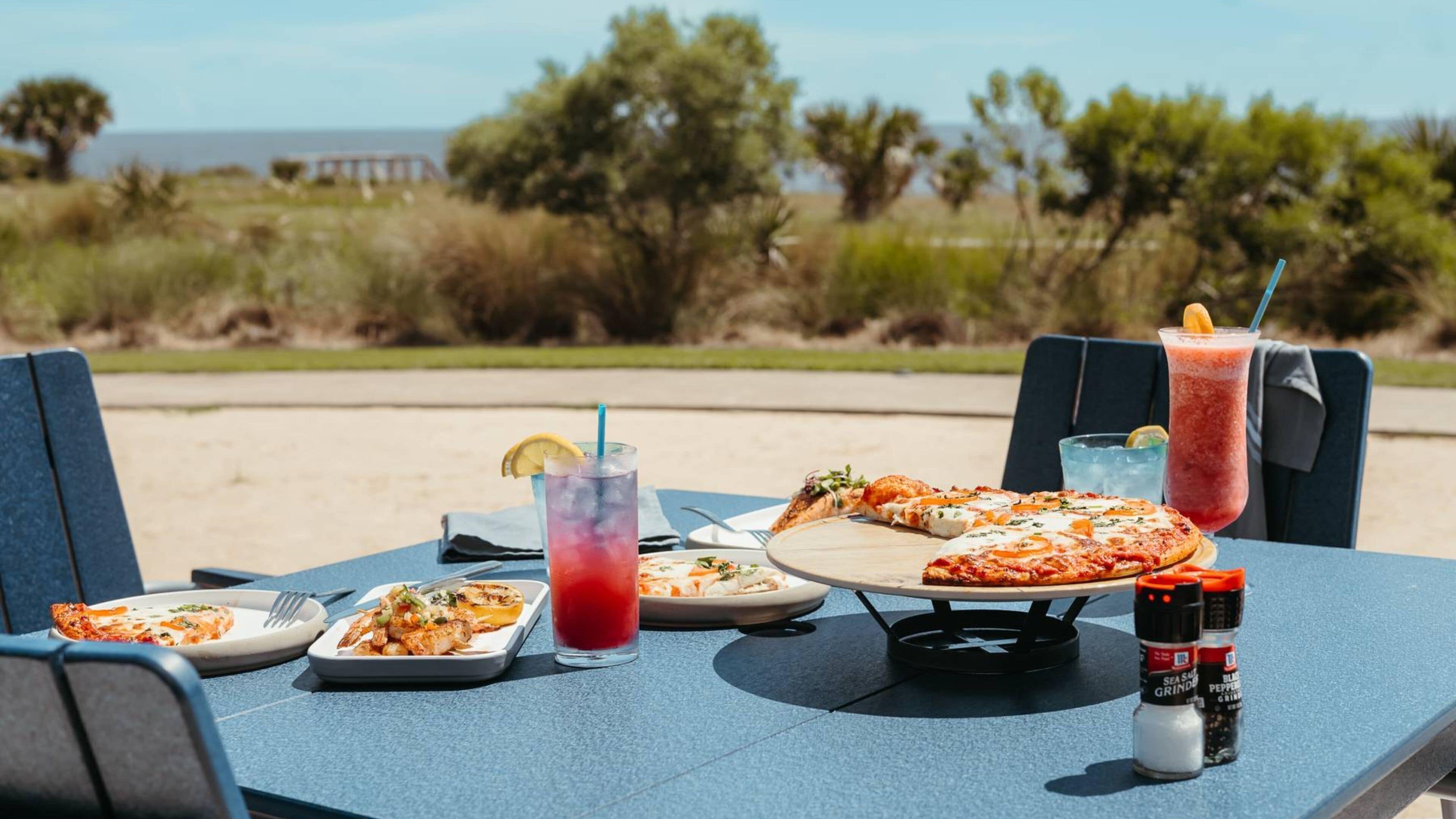 Una mesa con comida y bebidas en la playa, con sillas y vista al océano.