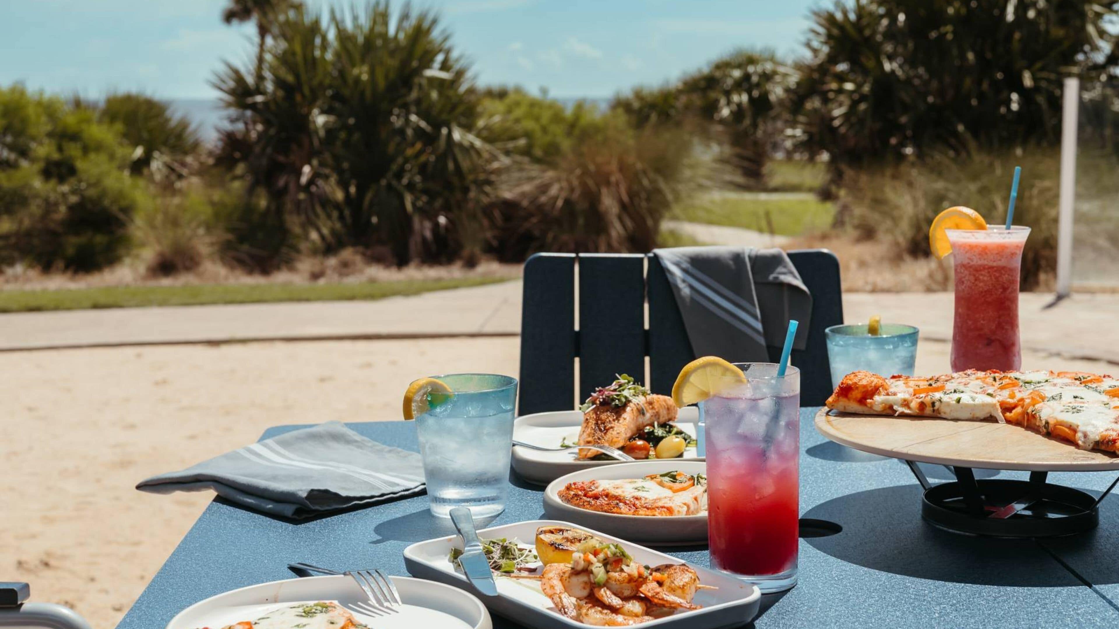 A table set outdoors with various dishes, drinks, and a pizza.