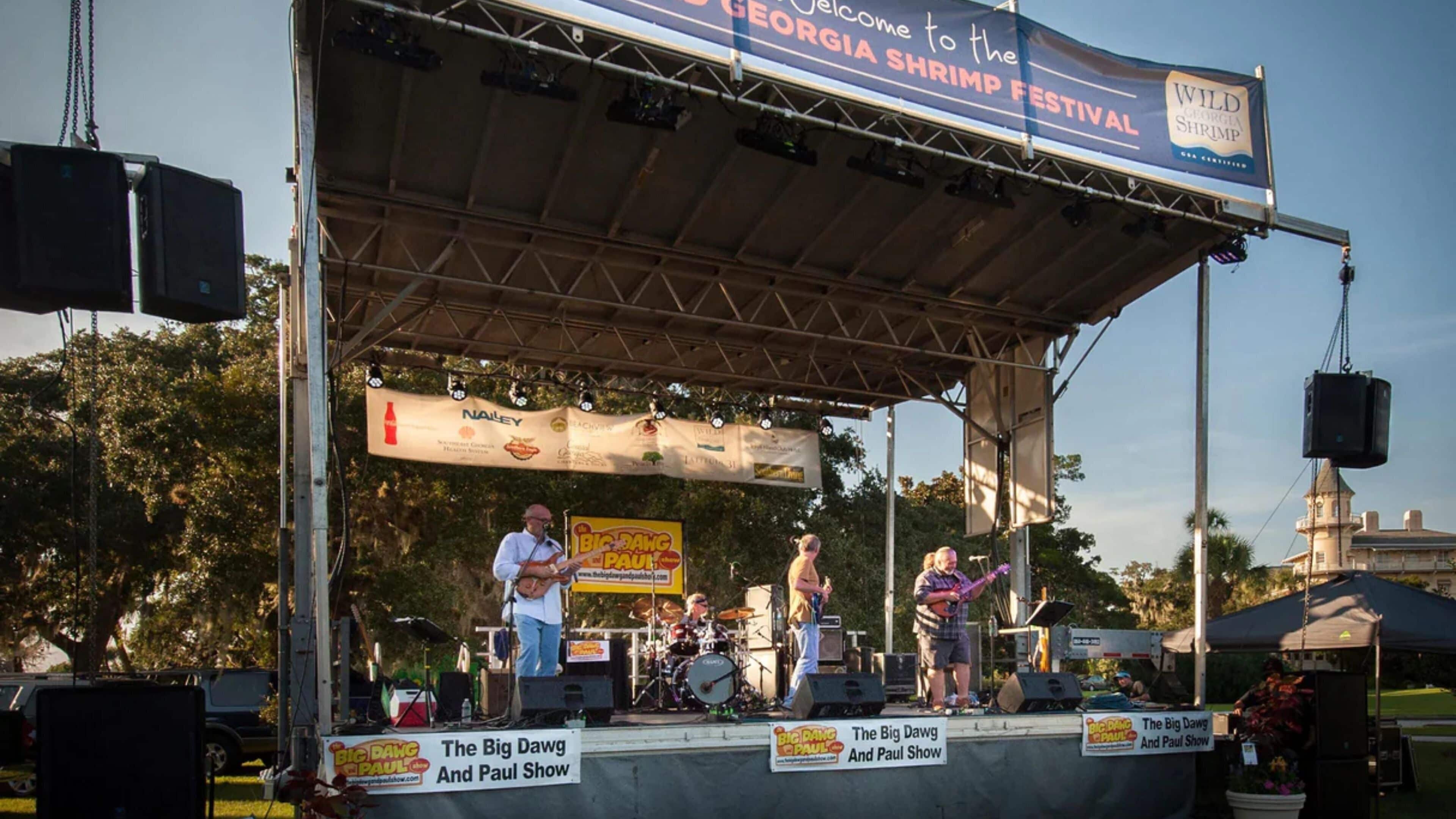 A stage with a band performing at an outdoor event, featuring banners for the Georgia Shrimp Festival and Big Dawg Paul Show.