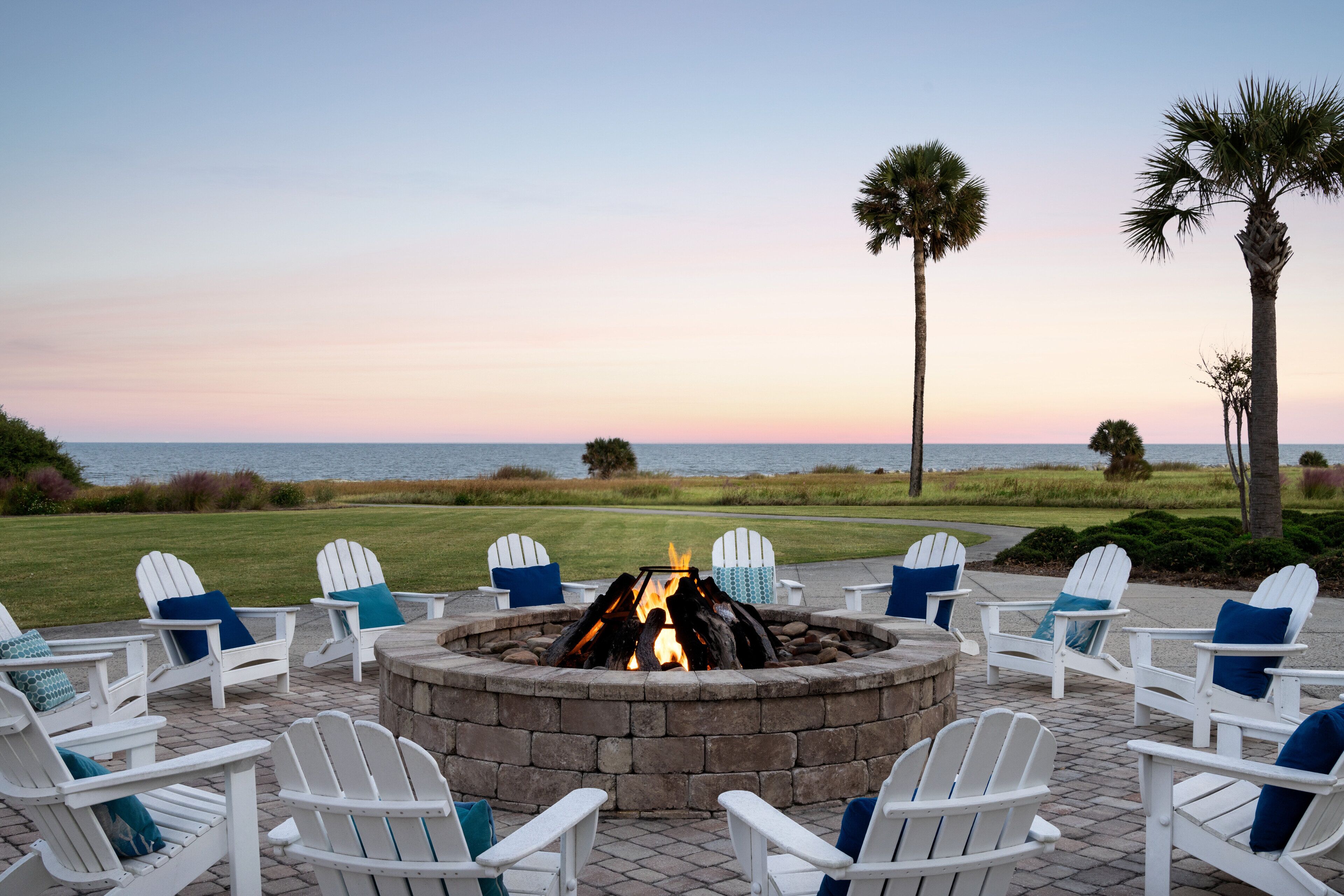 A fire pit surrounded by white Adirondack chairs with blue cushions, set on a patio with a view of the ocean and palm trees.