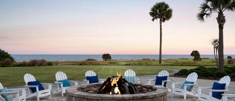 A fire pit surrounded by white Adirondack chairs with blue cushions, set on a patio with a view of the ocean and palm trees.
