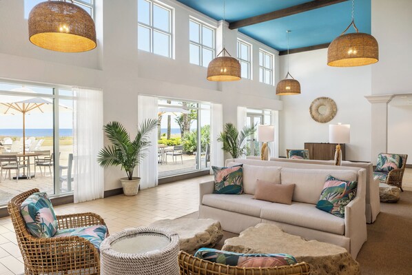 Amplia sala de estar con un sofá grande, sillas de mimbre y una mesa redonda. Hay ventanas y puertas grandes que dan a un patio exterior con vista al océano.