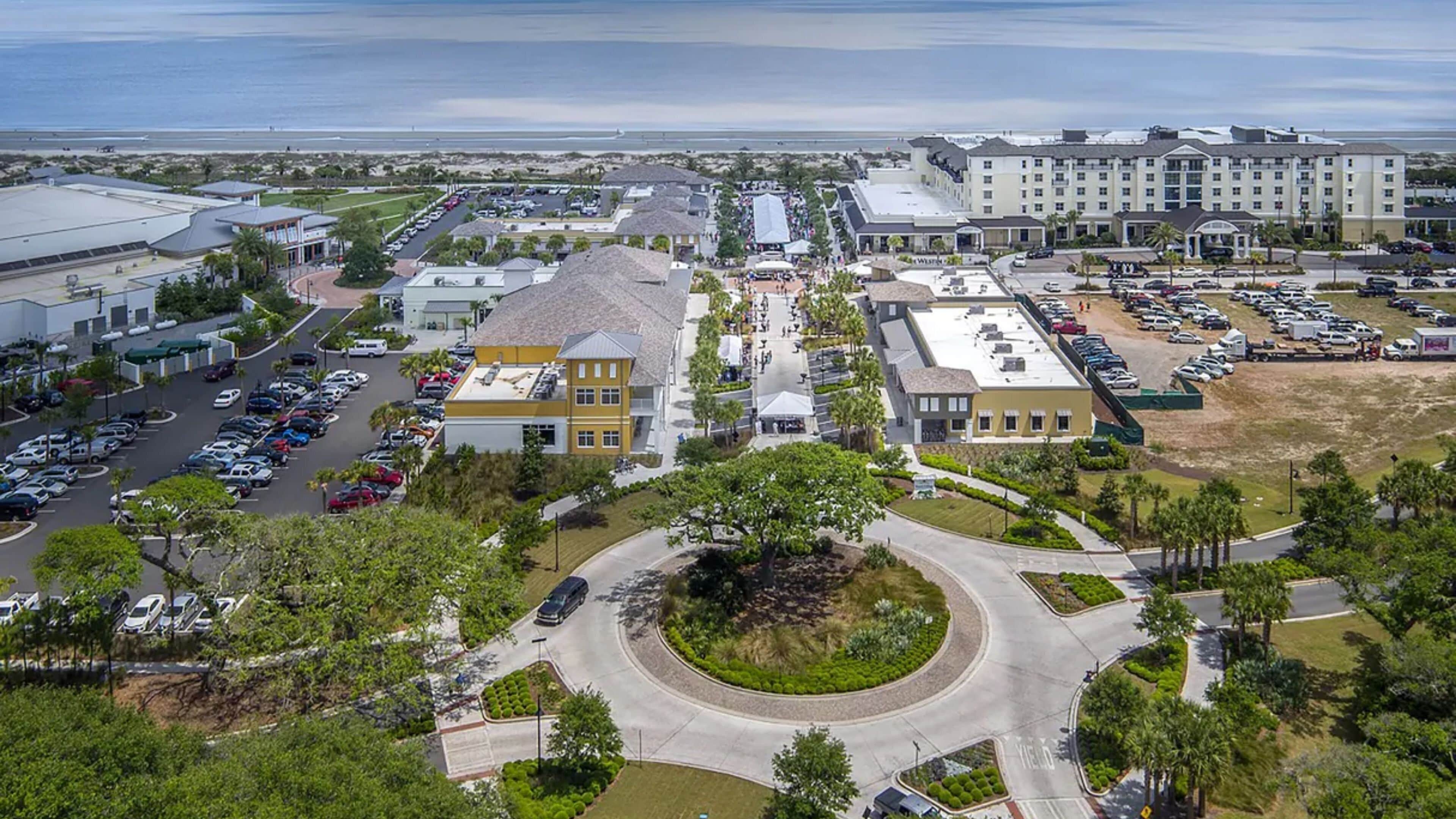 An aerial view of a coastal hotel area with multiple buildings, parking lots, and a roundabout.