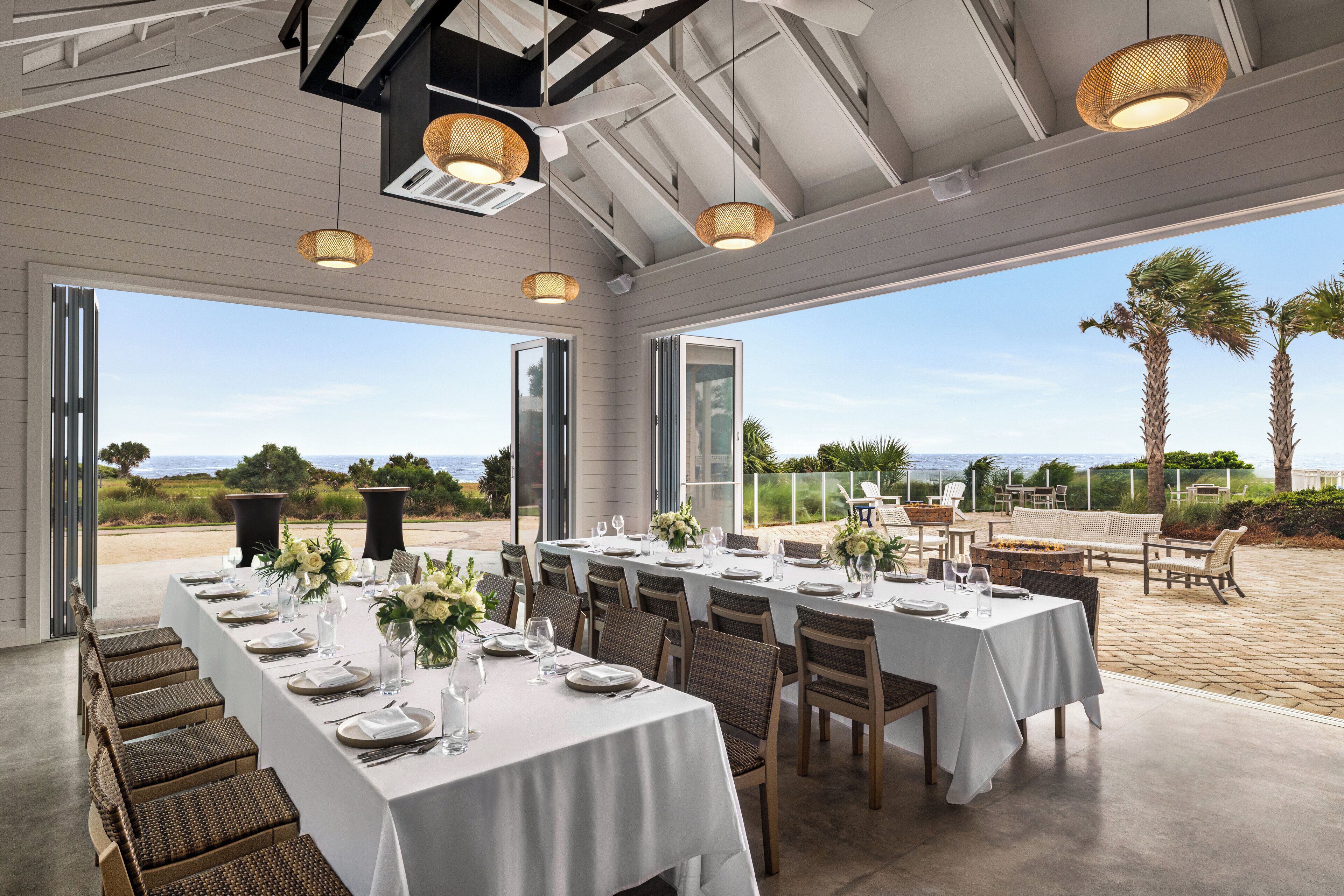 A long dining table set for a meal with chairs, a view of the ocean, and outdoor seating with a bench and chairs.
