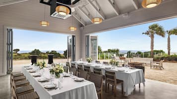Una larga mesa de comedor preparada para una comida, con sillas, vista al océano y asientos exteriores con una banca y sillas.