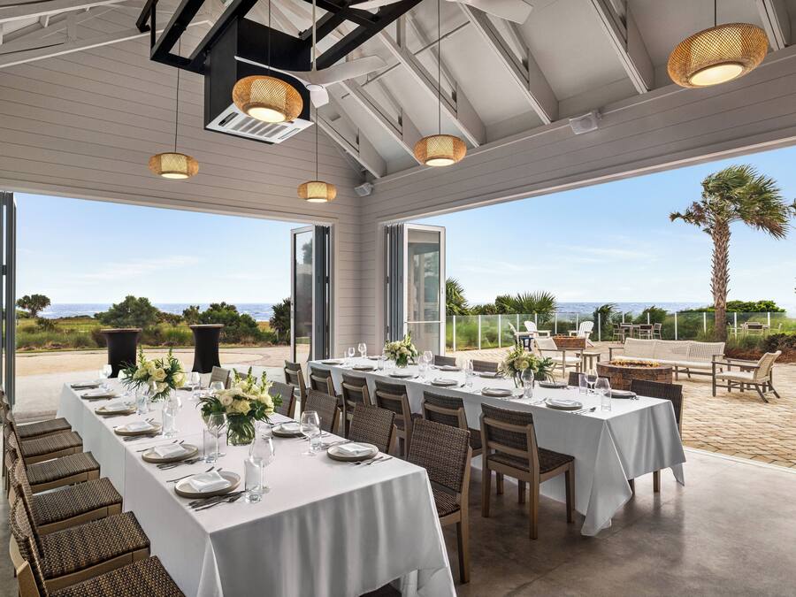 Una larga mesa de comedor preparada para una comida, con sillas, vista al océano y asientos exteriores con una banca y sillas.