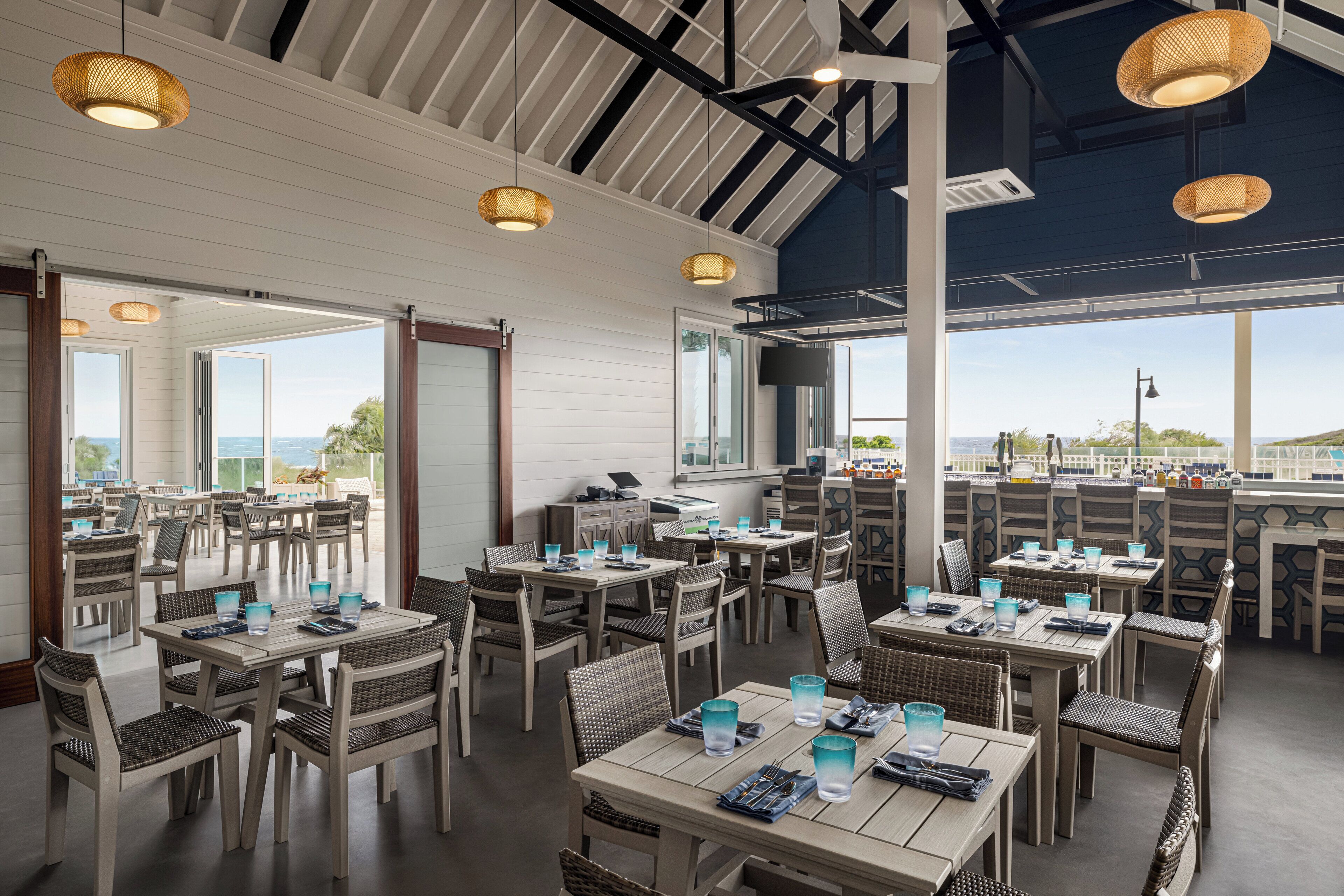 Un ristorante con diversi tavoli e sedie, ampie finestre che offrono vista sul mare e lampade in vimini appese.