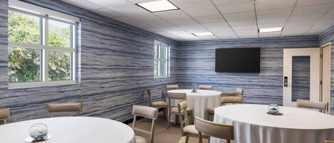 A conference room with round tables, beige chairs, a large window, and a flat-screen TV on the wall.