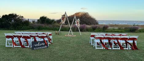 An outdoor wedding setup with white chairs and red sashes, a welcome sign, and a backdrop of the ocean.