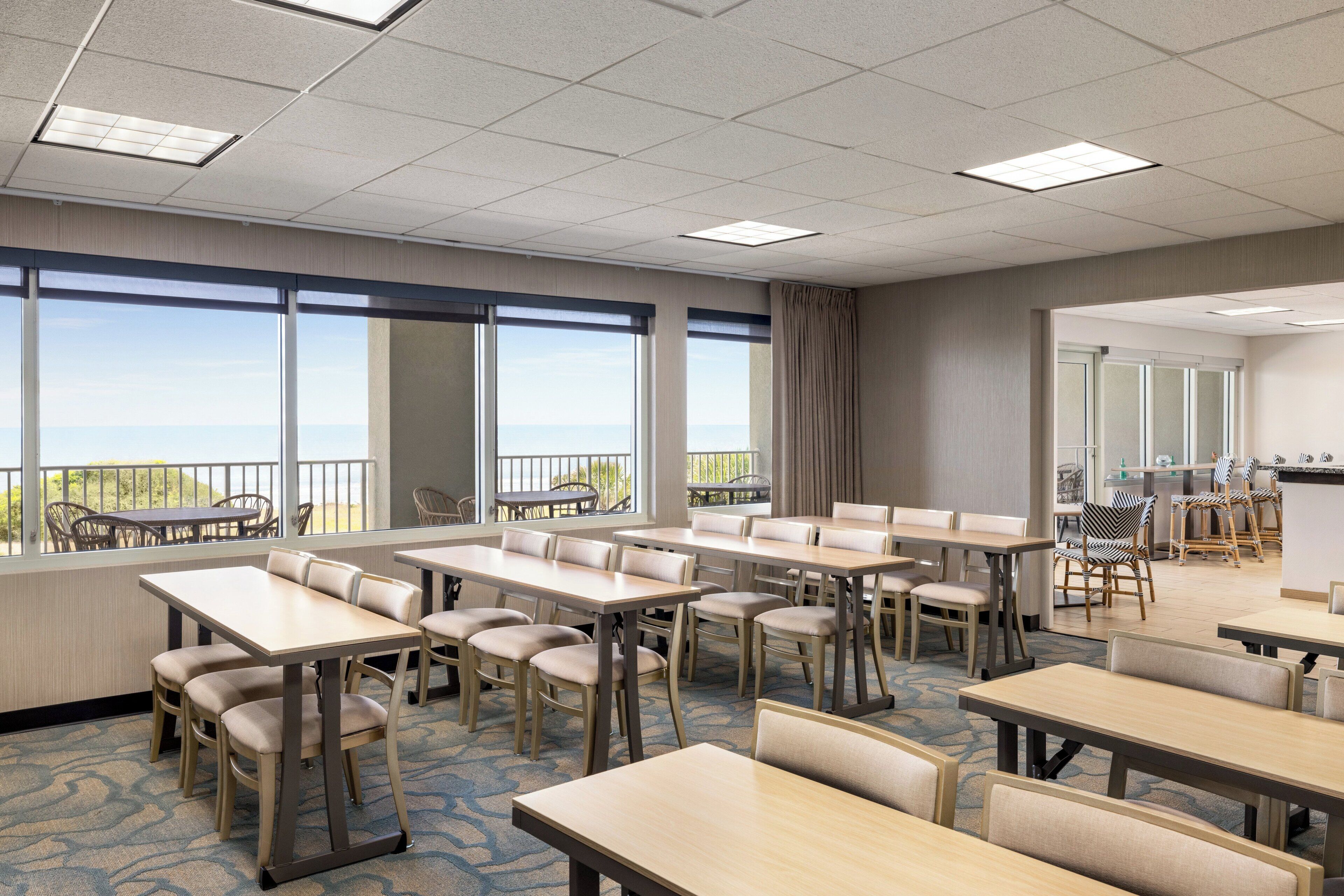 Un salón de conferencias con varias mesas y sillas, amplias ventanas con vistas y una zona de kitchenette.
