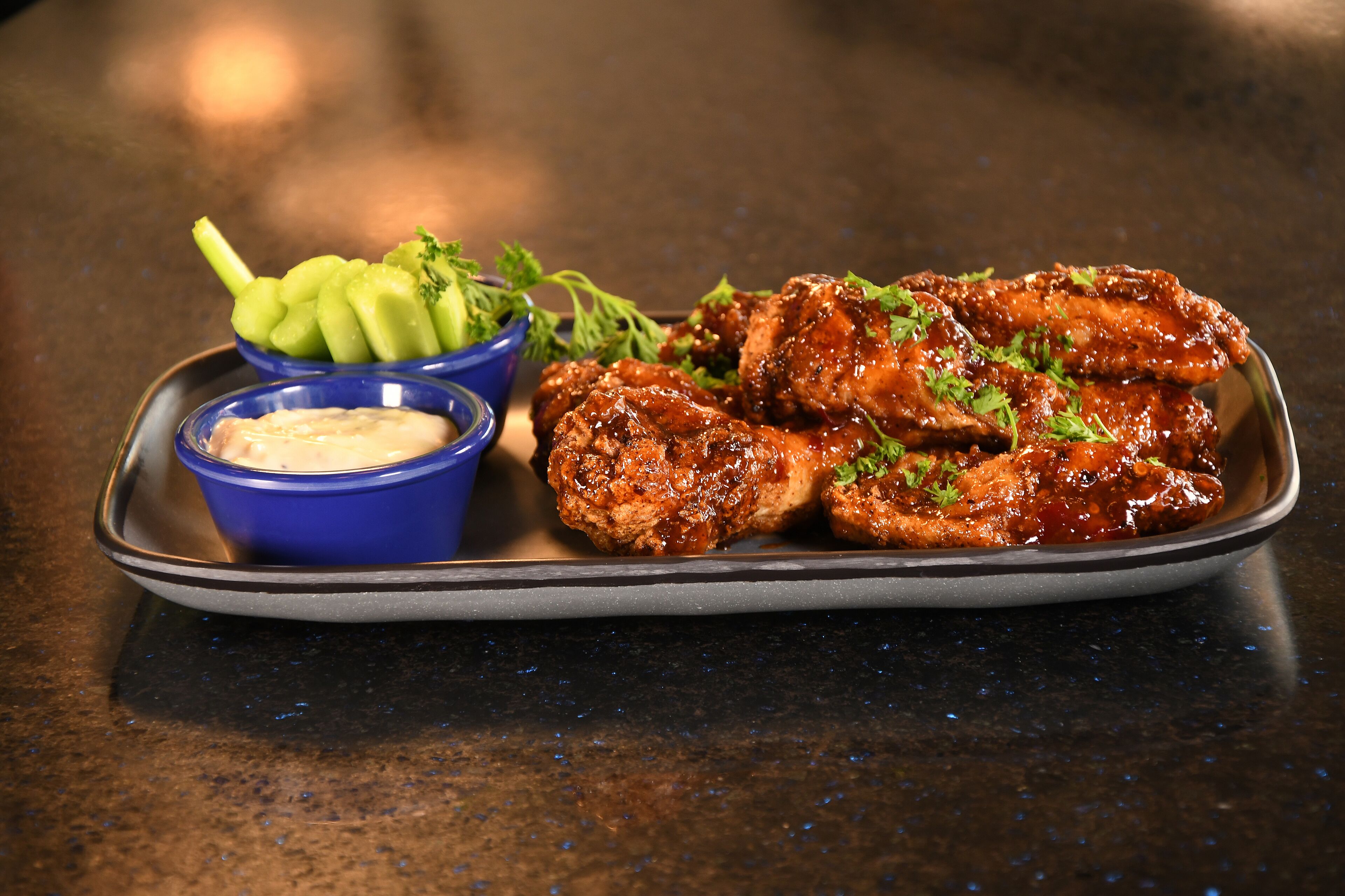 A plate of glazed chicken wings with a side of celery and a blue bowl of sauce.