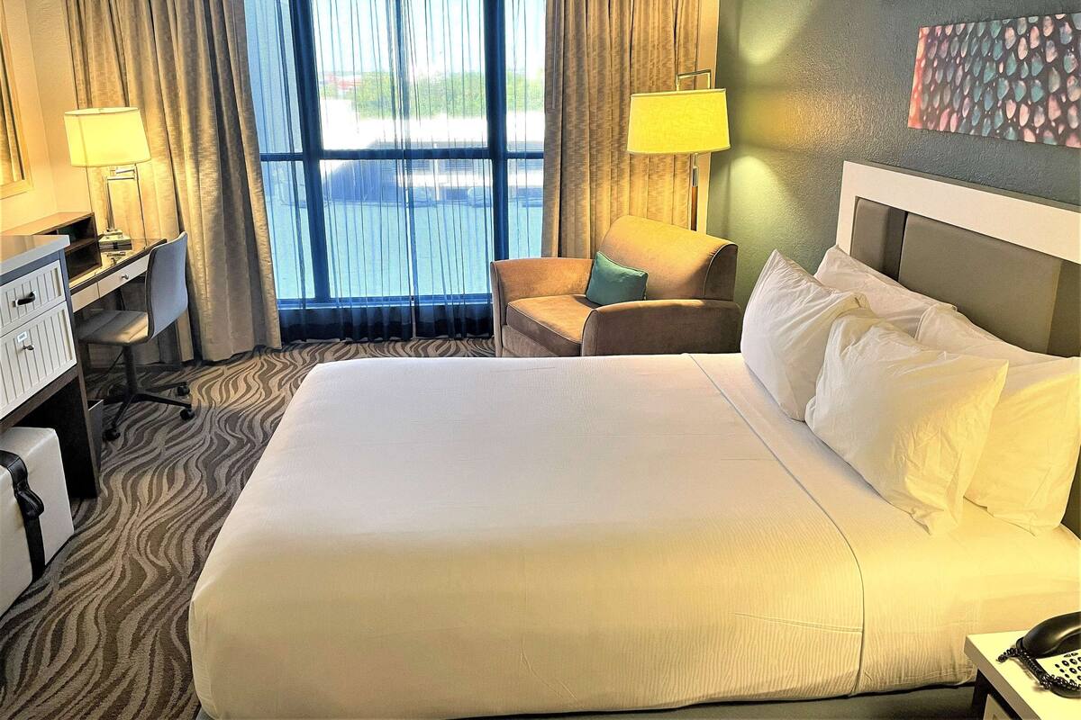 Ein Hotelzimmer mit einem großen Bett, einem Schreibtisch, einem Stuhl und Blick ins Freie durch das Fenster.