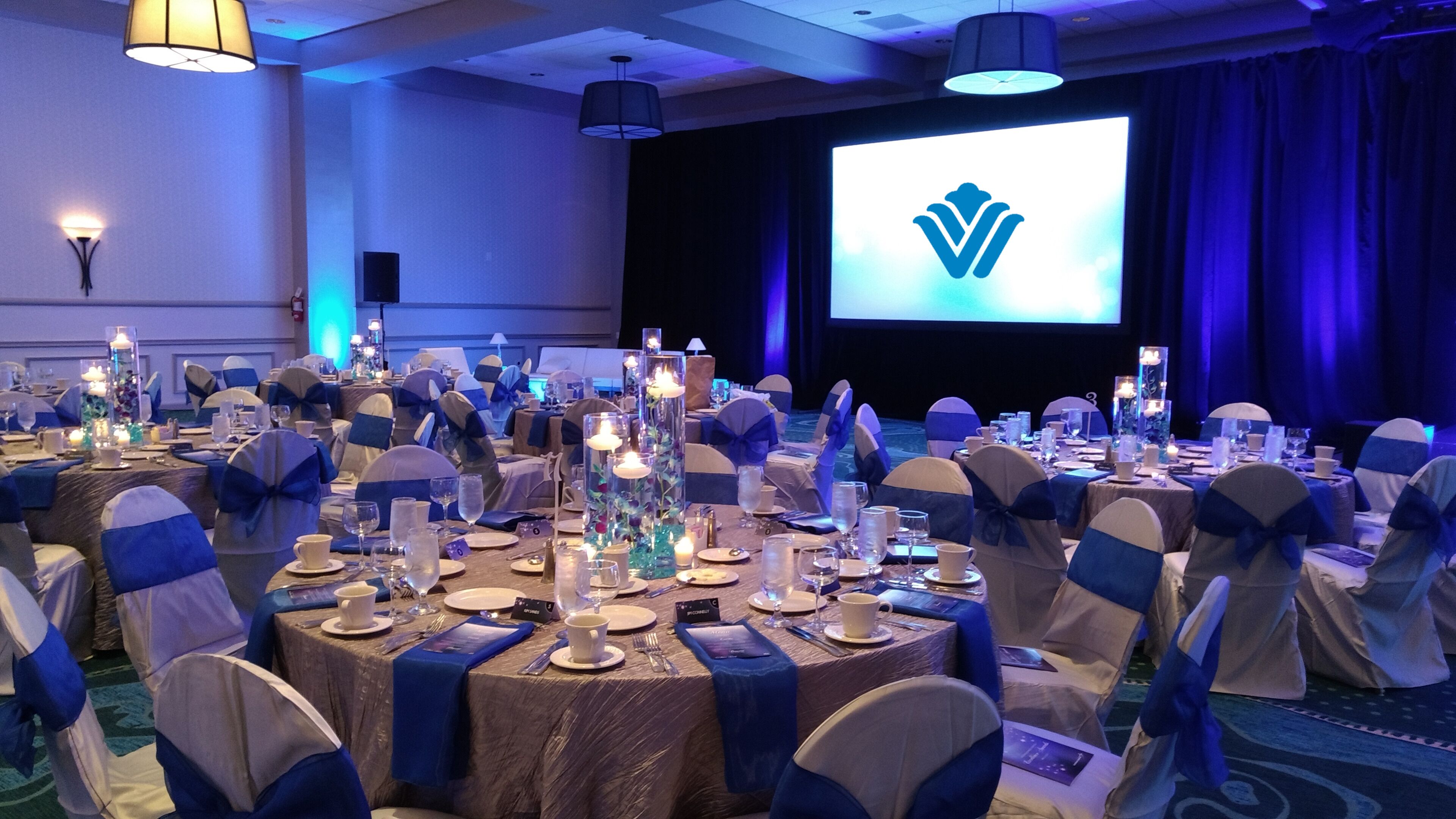 A banquet hall with round tables set for a formal event, featuring blue and white decor, candles, and a projection screen in the background.