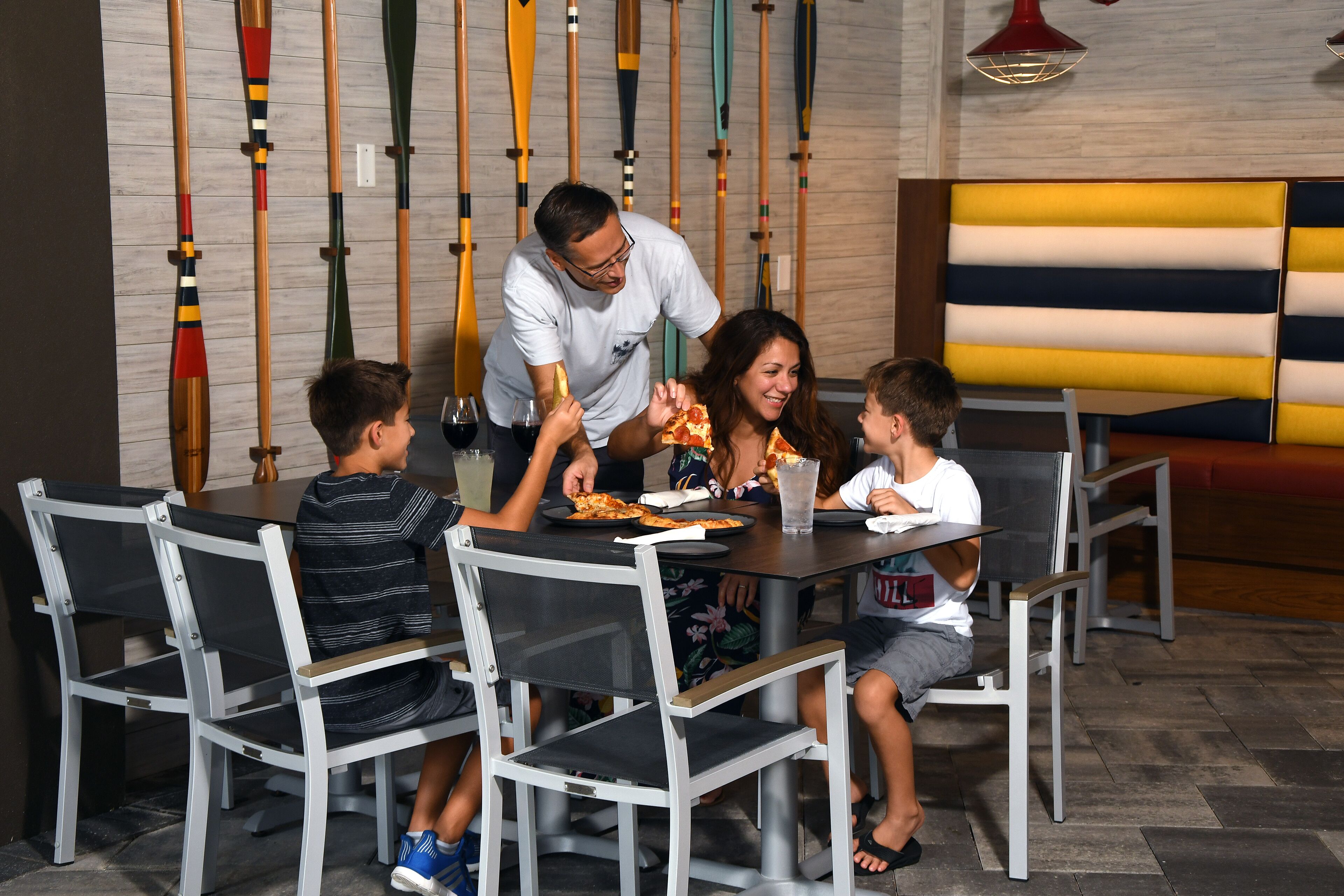 Una familia cenando en un restaurante con una decoración moderna, que incluye una pared con palas y un área con asientos tipo cabina de colores.