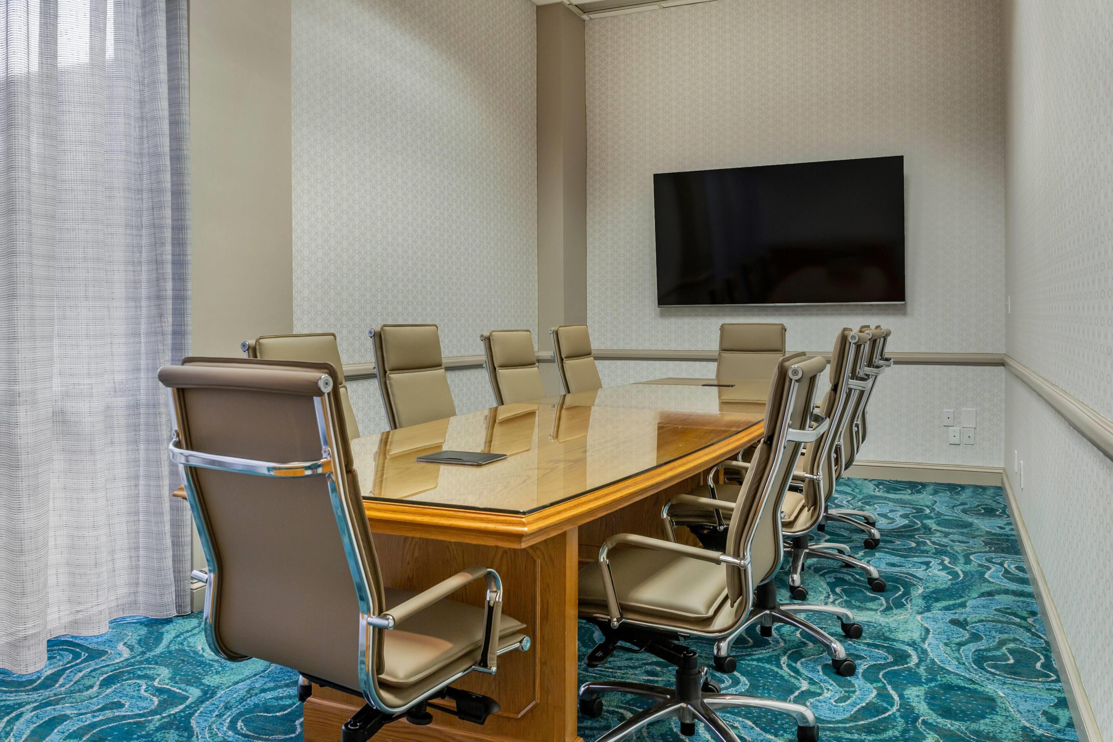 A conference room with a glass table, beige chairs, a mounted TV, and patterned carpet.