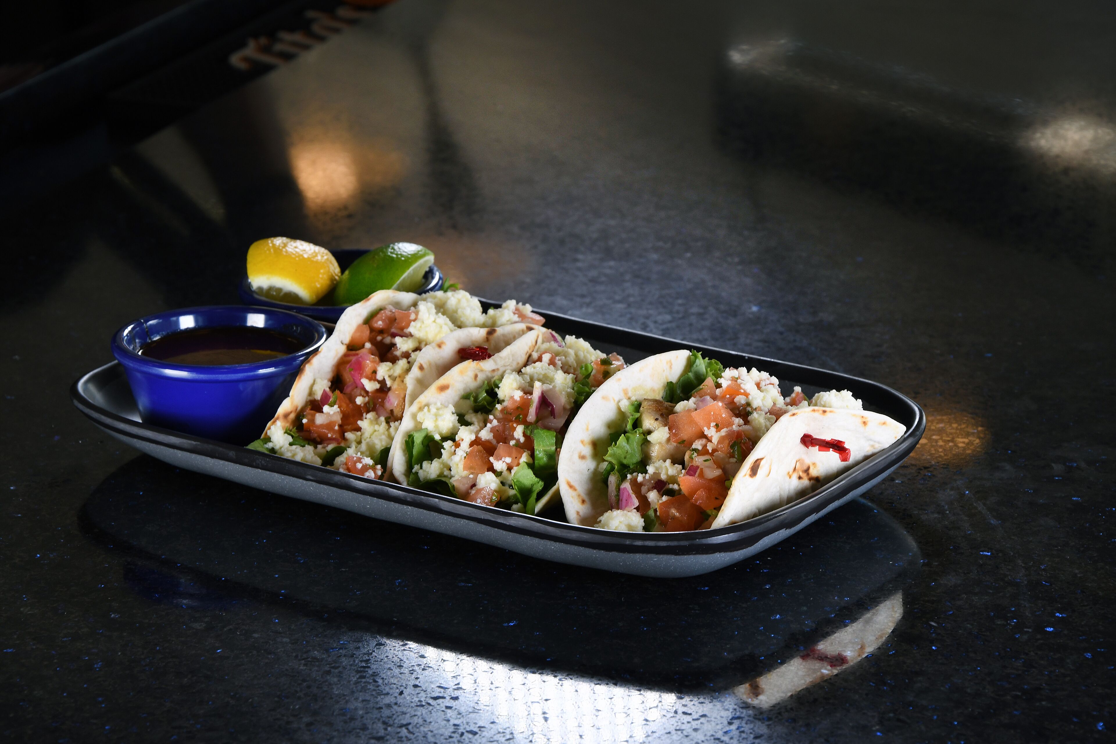 A tray with three tacos, a small bowl of sauce, and a lime on a dark countertop.