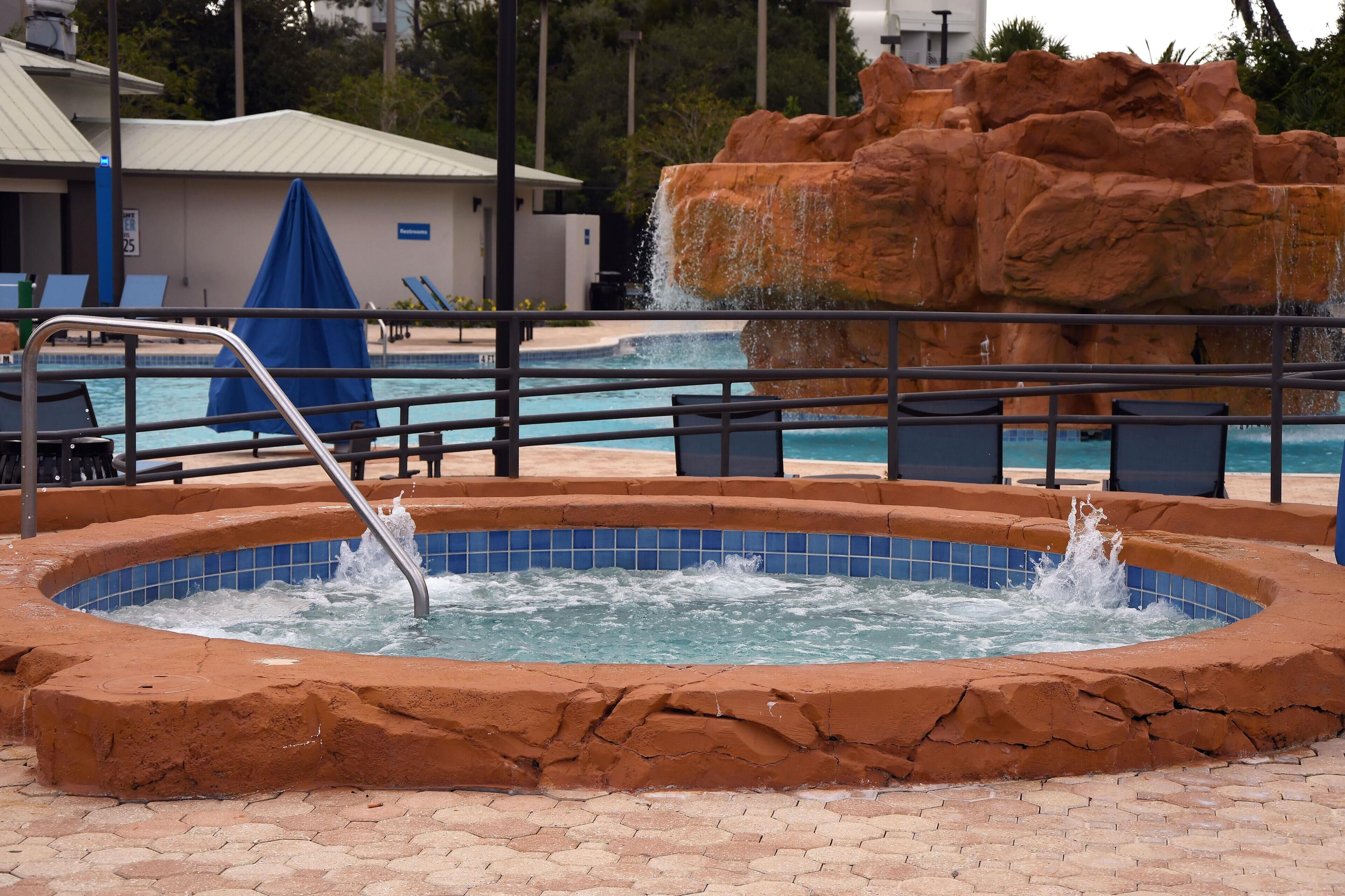A hot tub with a rocky backdrop, a pool with lounge chairs, and a building with a sign.