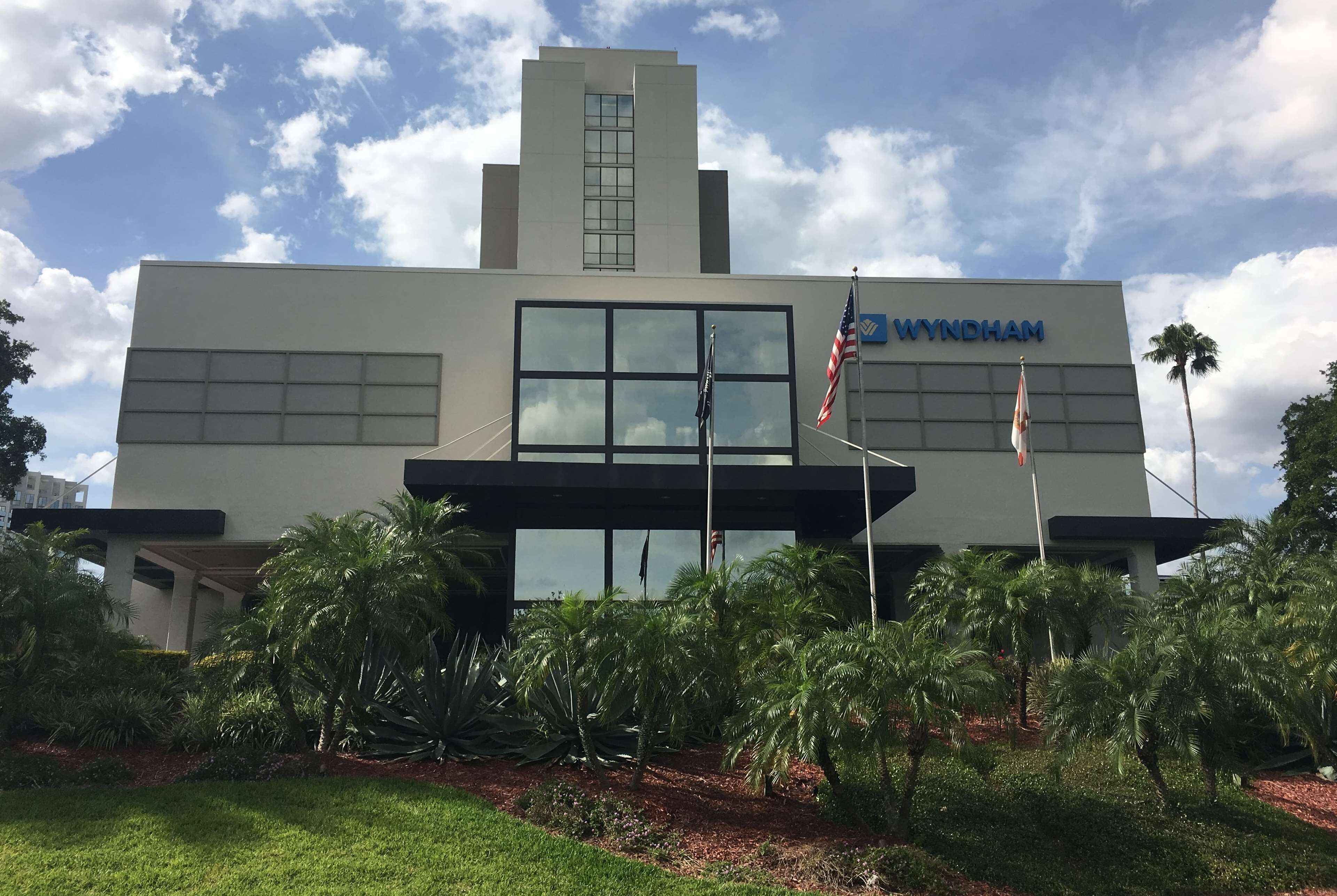A modern building with the name 'Wyndham' on the facade, featuring large windows, a flagpole with the American flag, and a well-maintained garden with palm trees.