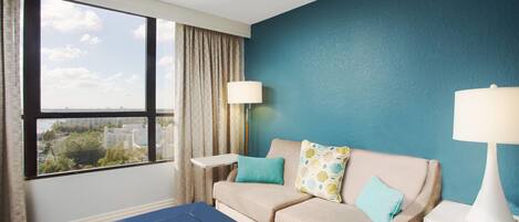 A hotel room with a sofa, a small table, a lamp, and a view of the city through large windows.