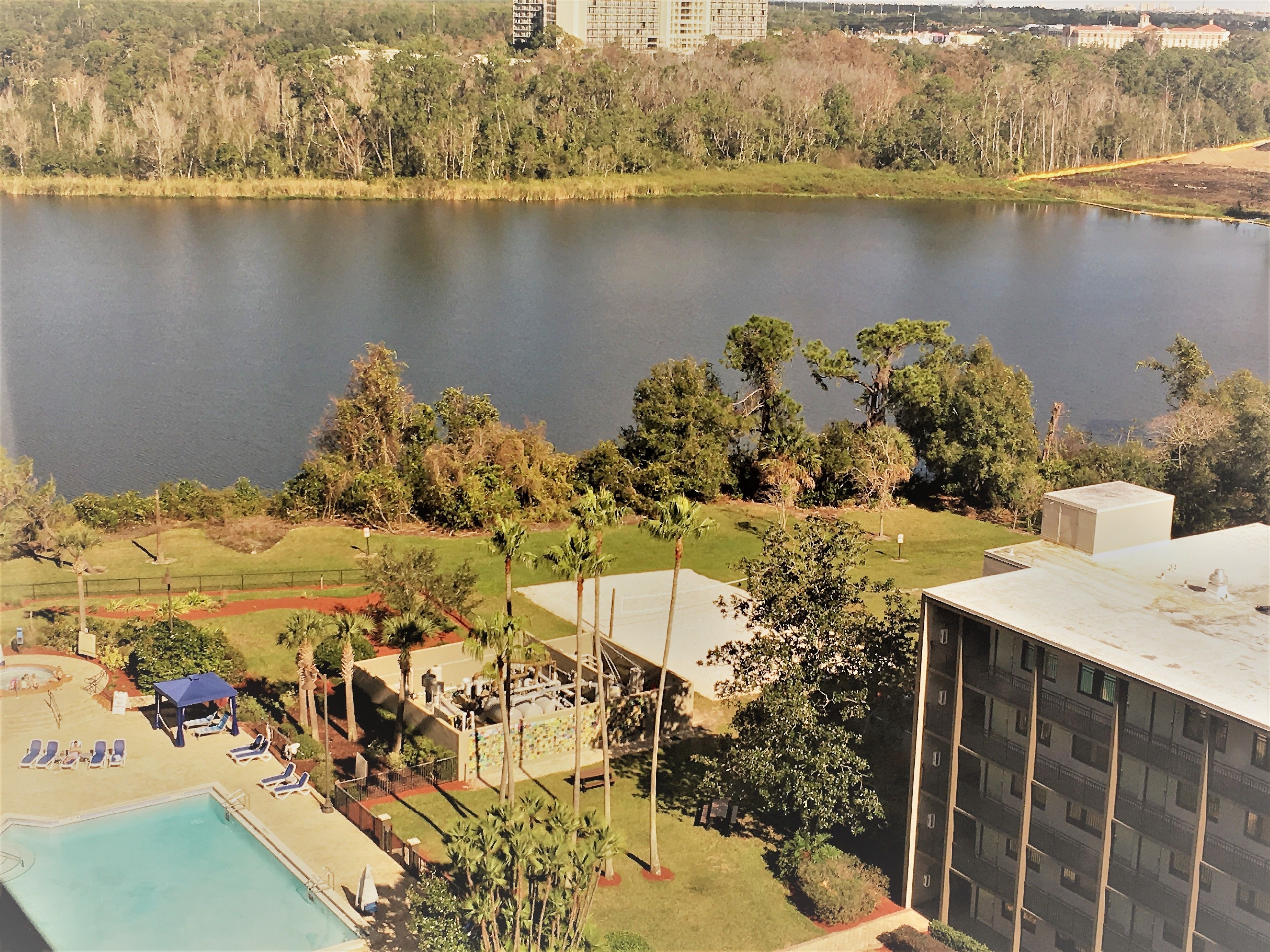 A view of a hotel with a swimming pool, a tennis court, and a large body of water in the background.
