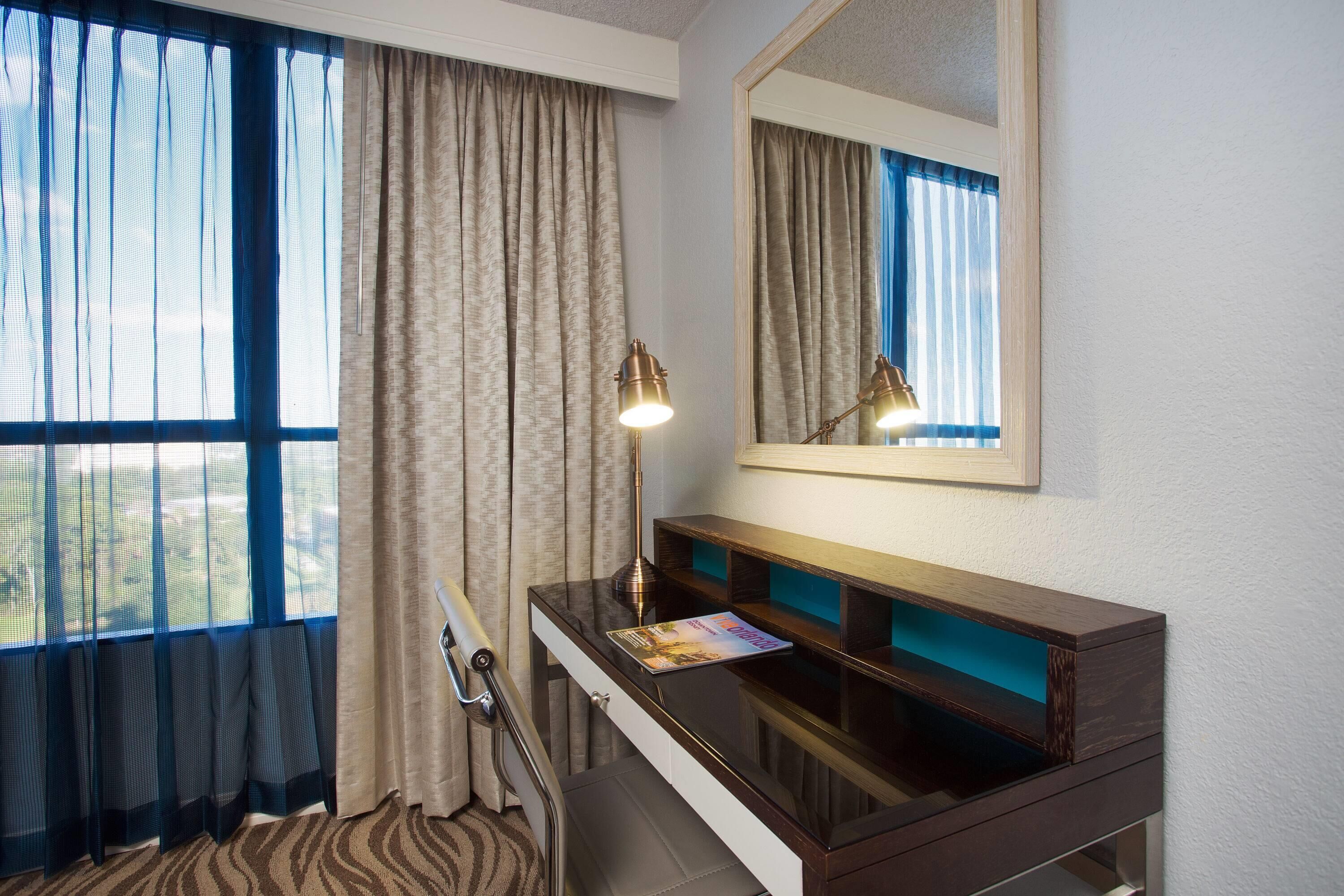 A hotel room with a desk, a mirror, a lamp, and a window with curtains.