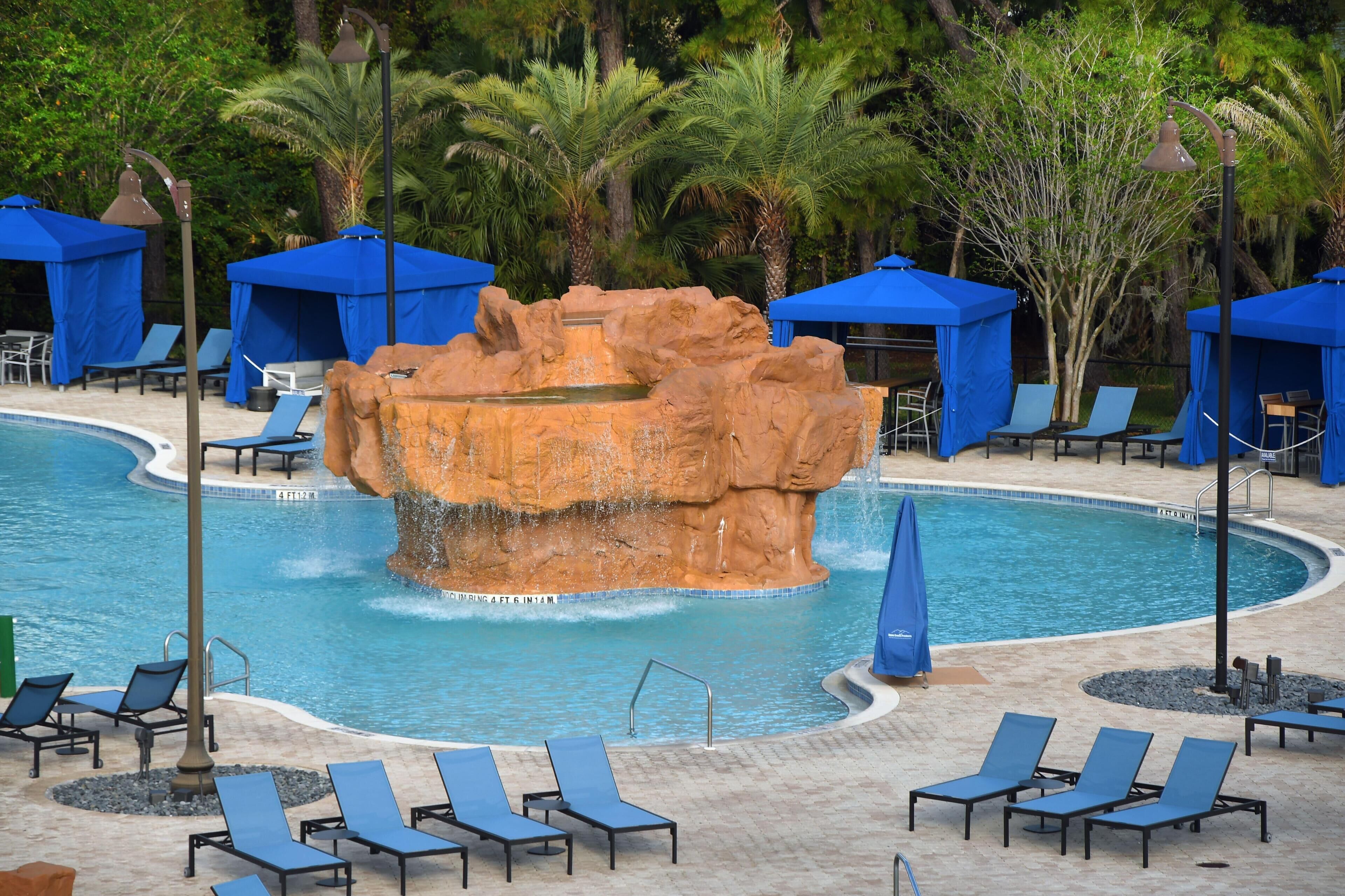 Zona de piscina con sombrillas azules, sillas reclinables y una formación rocosa en el centro.