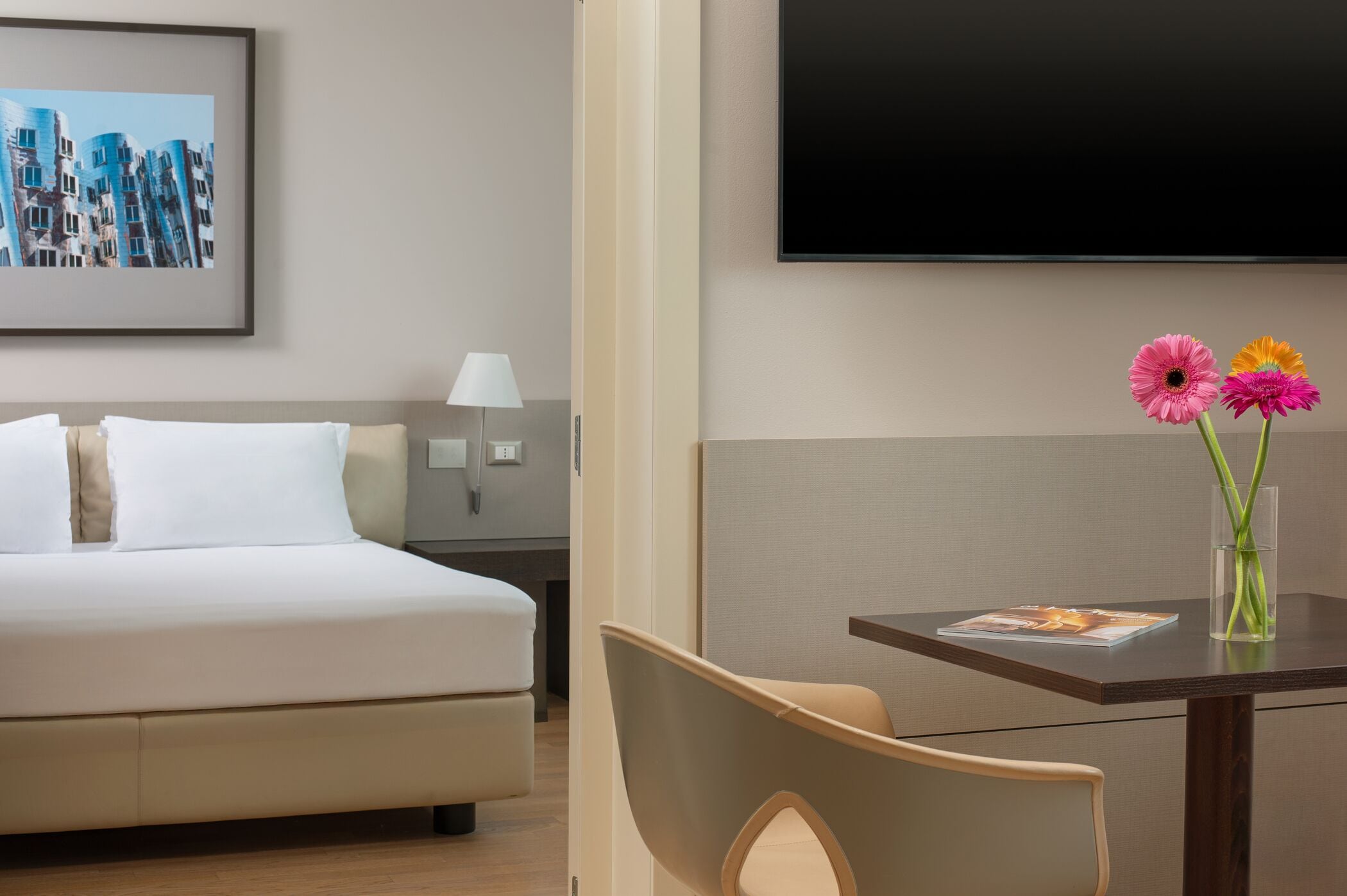 A hotel room with a bed, a nightstand with a lamp, a TV, and a table with a magazine and flowers.