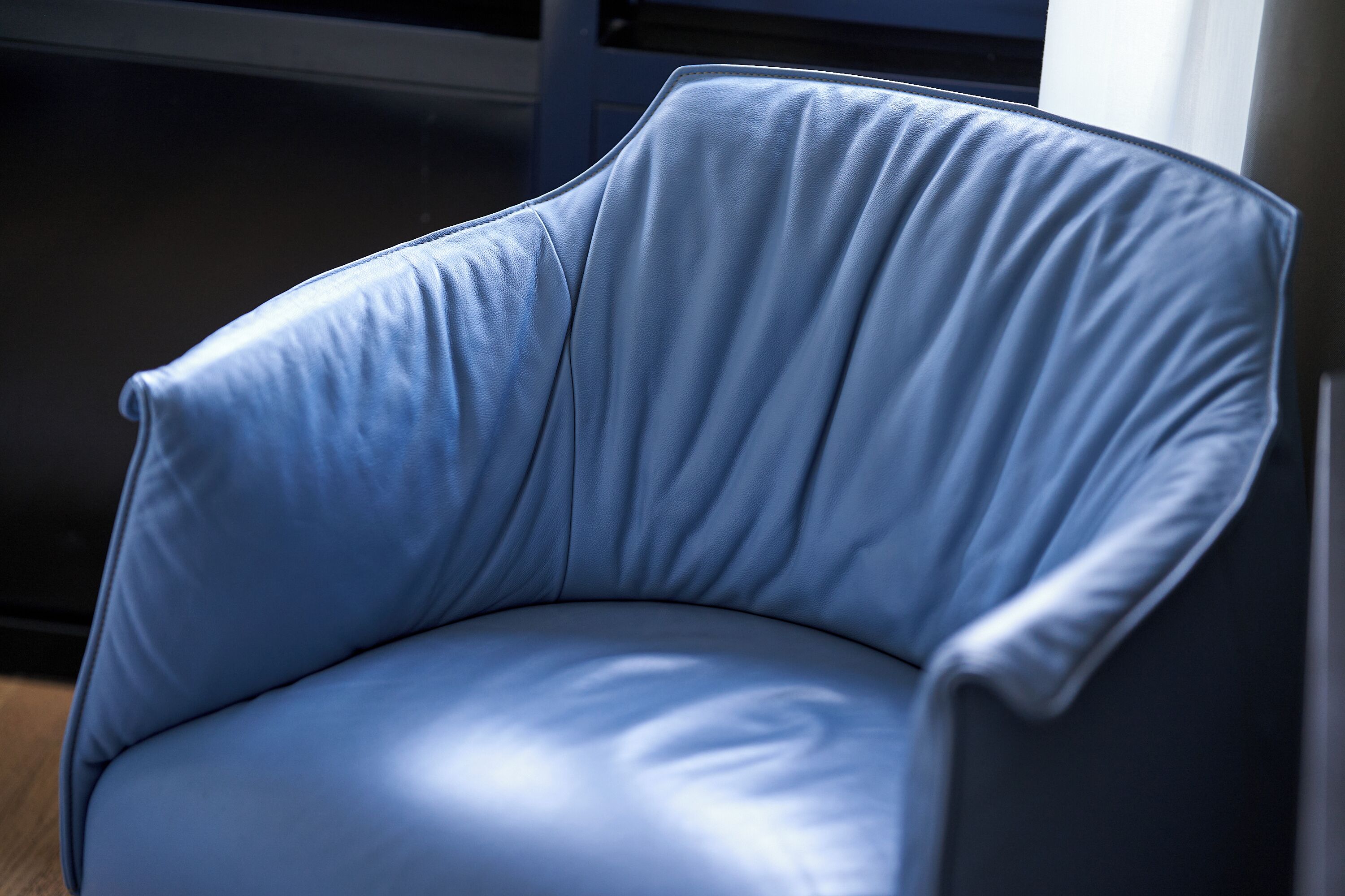 Uma poltrona de couro azul com encosto e apoio de braço curvos, colocada em um quarto com chão escuro e uma janela ao fundo.