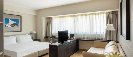 A modern hotel room with a bed, a sofa, a desk with a TV, and a large window with curtains.