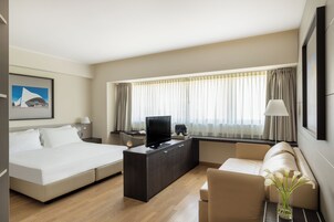 Una habitación de hotel moderna con cama, sofá, escritorio con televisor y un amplio ventanal con cortinas.
