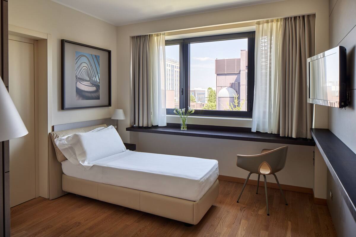 A hotel room with a bed, a desk, a chair, a TV, a window with curtains, and a framed picture on the wall.