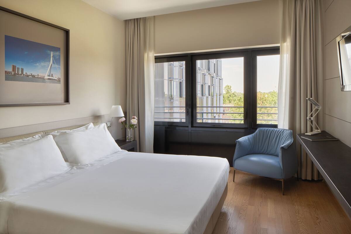 A hotel room with a large bed, a chair, a desk, and a window with a view of buildings.