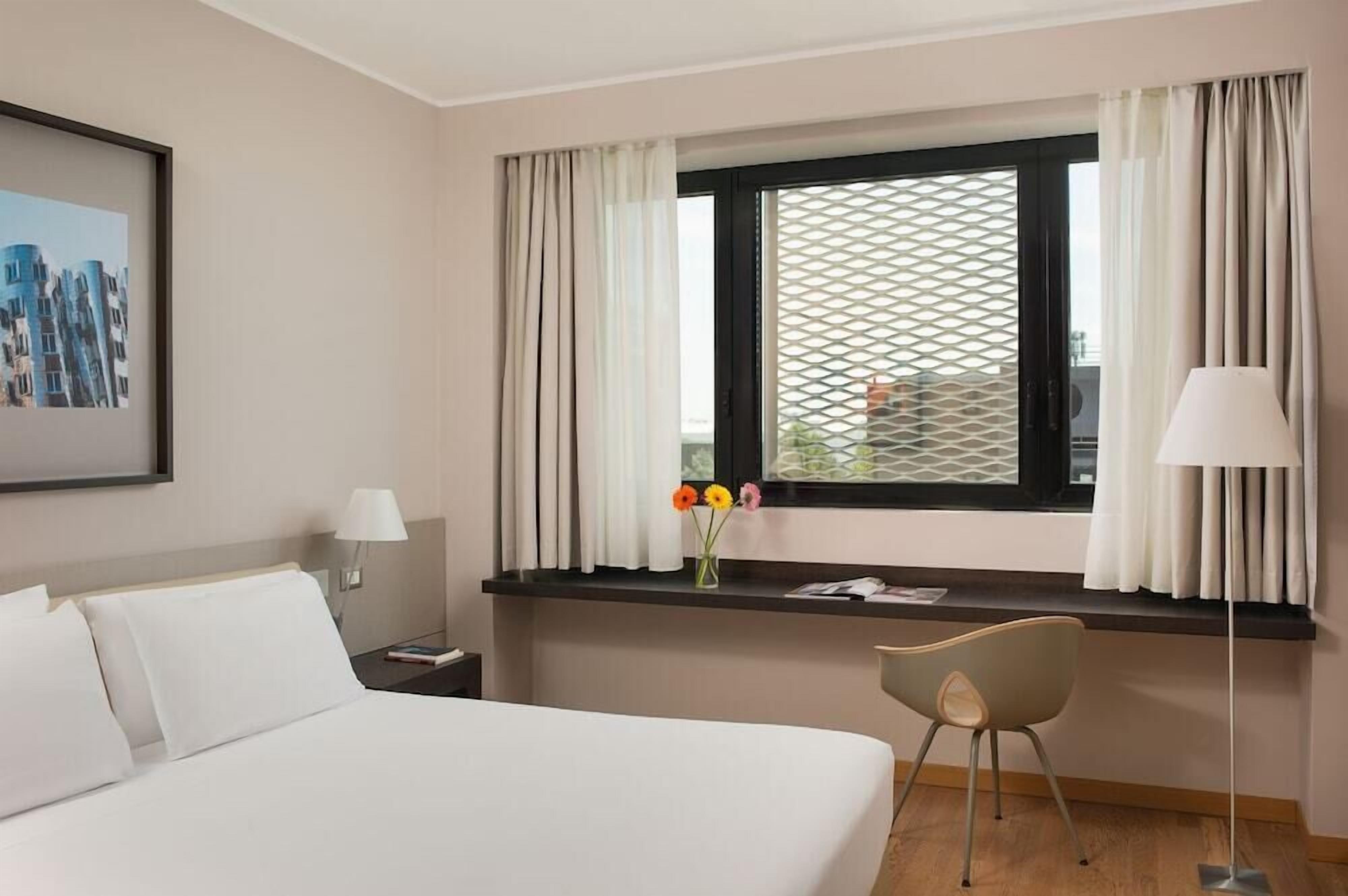 Habitación de hotel con cama, escritorio y silla, ventana con cortinas, lámpara y un cuadro en la pared.