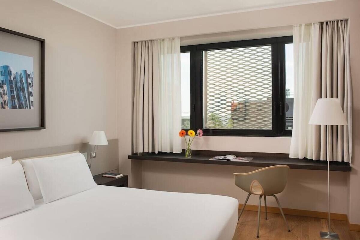 Une chambre d’hôtel avec un lit, un bureau et une chaise, une fenêtre avec des rideaux, une lampe et un tableau encadré au mur.