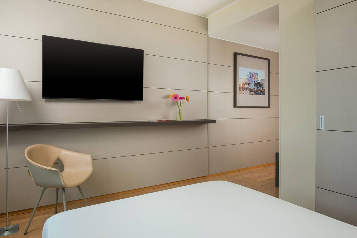 A modern hotel room with a flat-screen TV, a framed picture on the wall, a small table with a vase of flowers, a chair, and a bed.