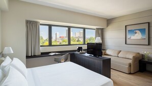 Hotelkamer met een bed, bureau, televisie, bank en een groot raam met uitzicht op de stad.