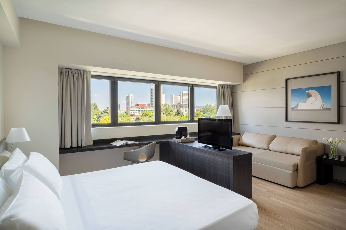 A hotel room with a bed, a desk, a TV, a sofa, and a large window with a city view.