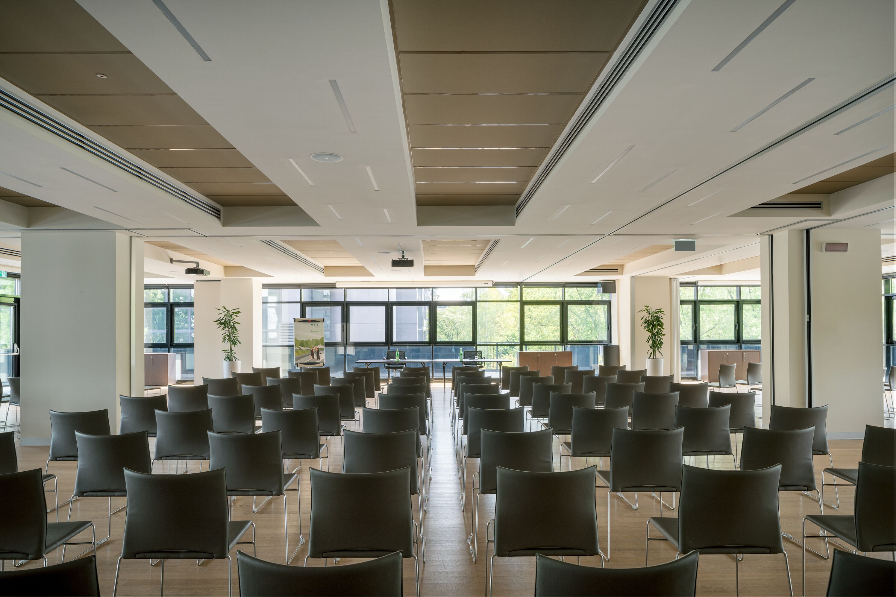Un salón de conferencias con filas de sillas orientadas hacia una zona de presentaciones, ventanas amplias y rejillas de ventilación en el techo.