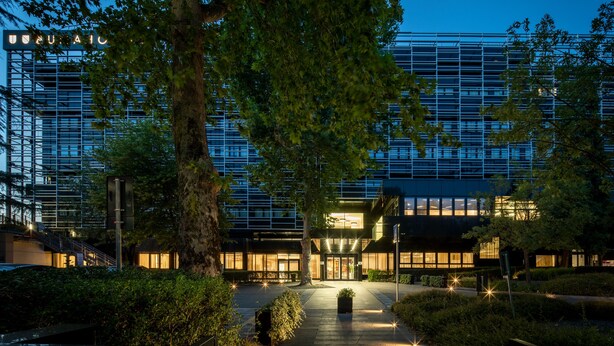 Een modern gebouw met een glazen gevel, omringd door bomen en een goed verlicht entreegebied.