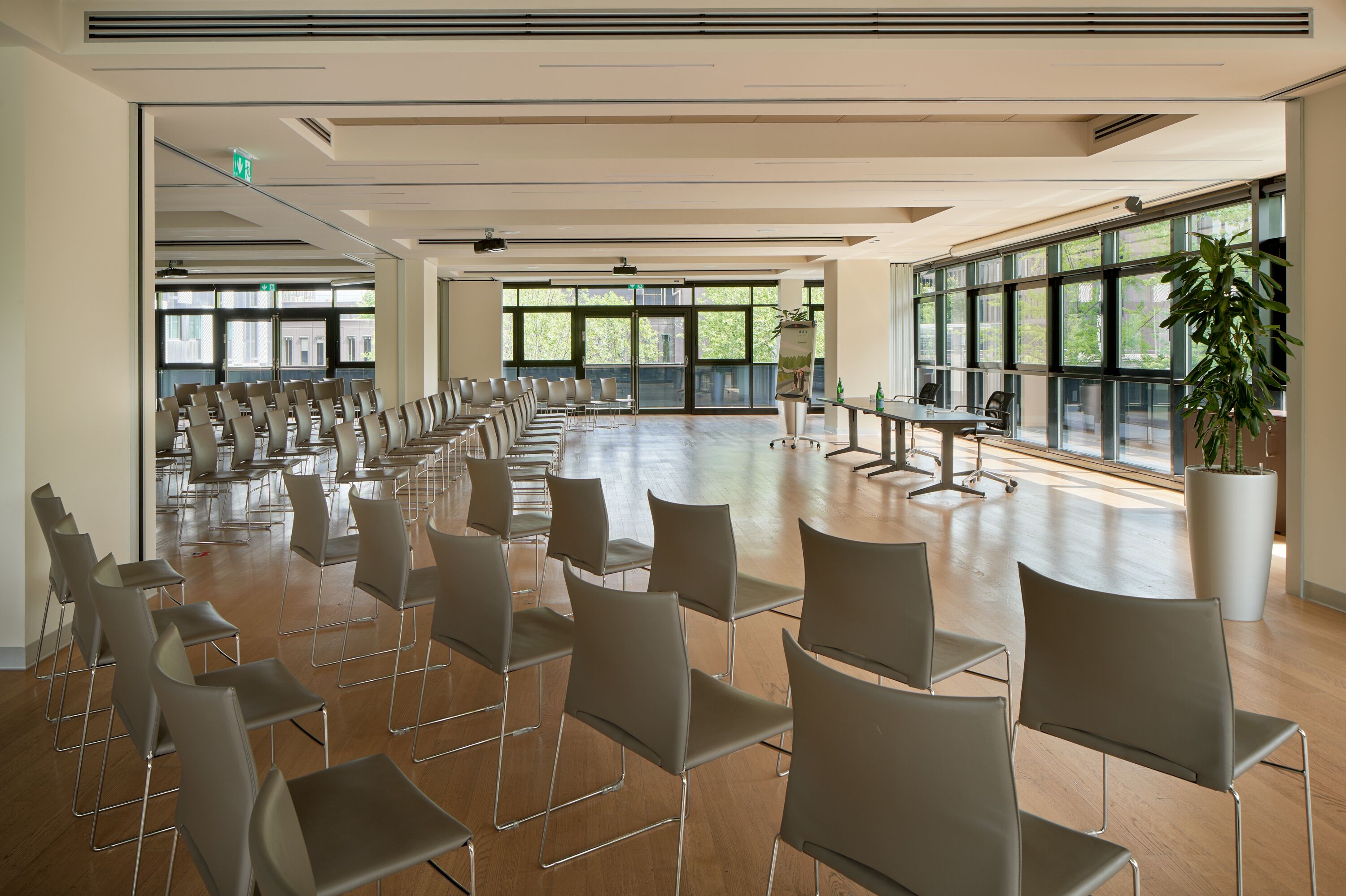 Uma sala de conferências ampla, com fileiras de cadeiras, grandes janelas e um vaso de plantas.
