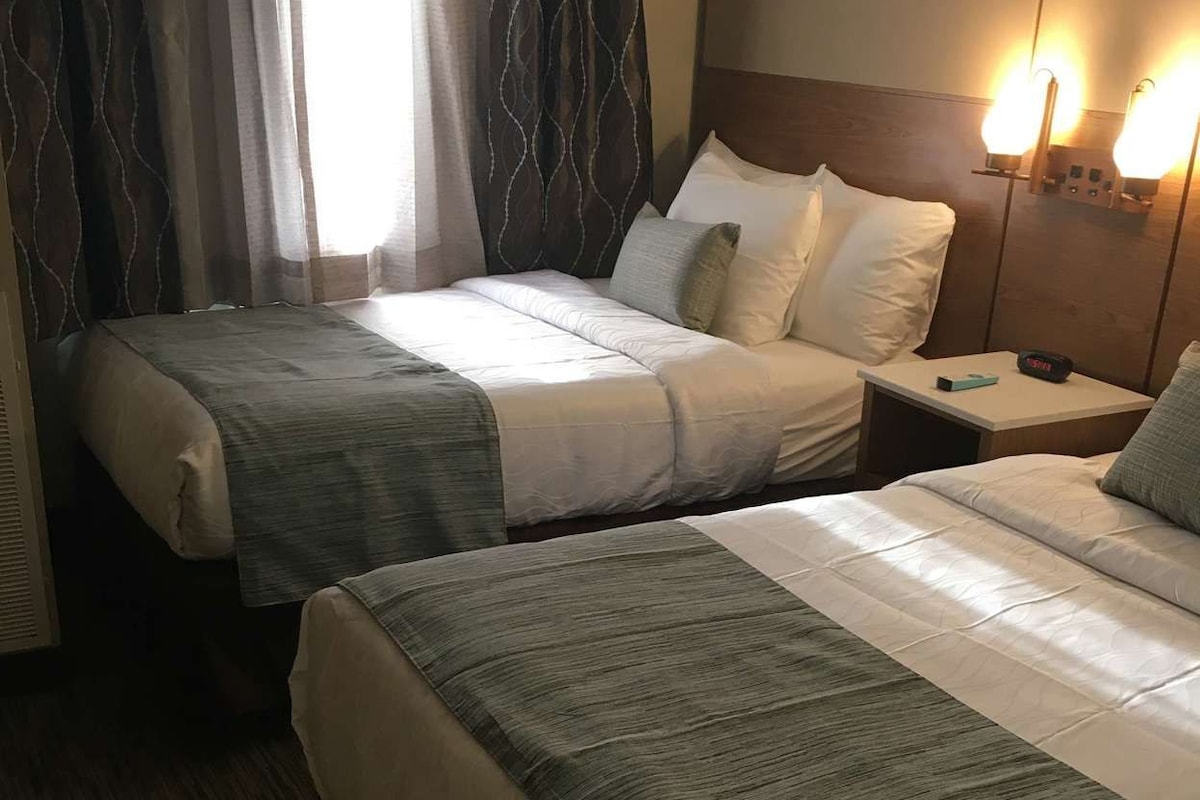 Habitación de hotel con dos camas, una mesita de noche, una lámpara y una ventana con cortinas.