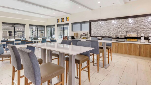 A modern dining area with high tables and chairs, a kitchen counter with appliances, and large windows providing natural light.